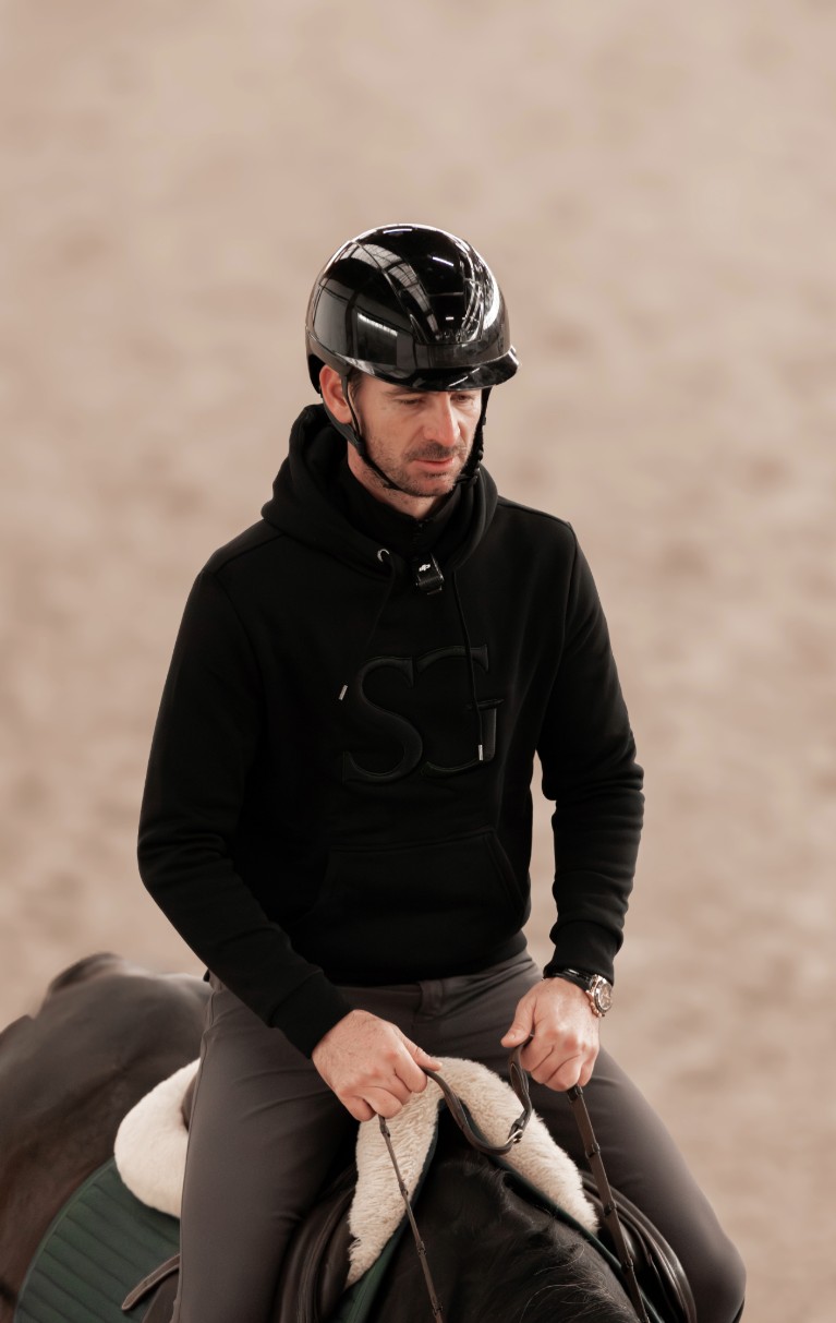 Cavalier en hoodie noir et casque noir, assis sur un cheval, en intérieur, tenue décontractée pour équitation.