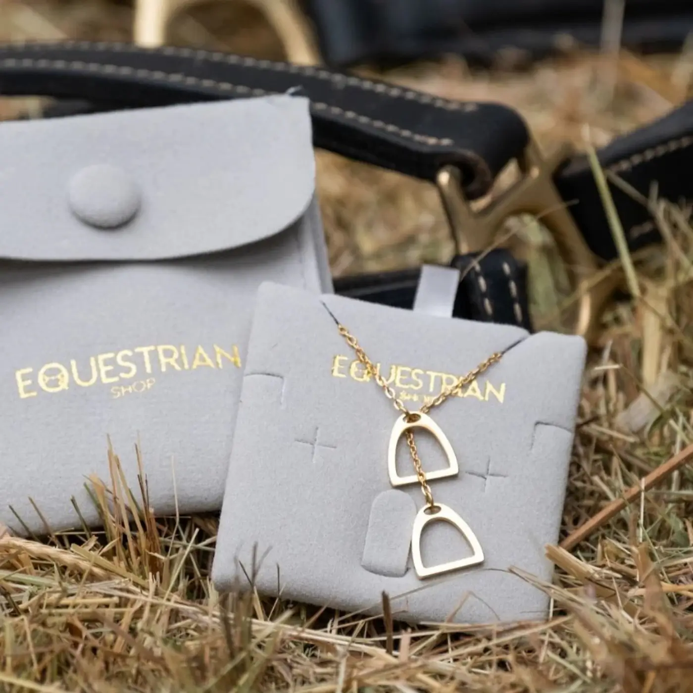 Collier doré à maillons mi-larges avec pendentifs en forme d’arche, posé sur support gris embossé "EQUESTRIAN SHOP" près d’un harnais noir, en herbe.